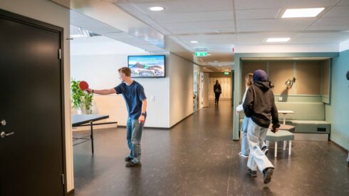 Skolkorridor som elever går i och änden av ett pingisbord där en elev står med pingisrack och servar en boll
